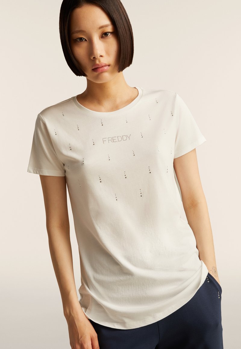 Camiseta blanca de manga corta con la palabra "FREDDY" en tachuelas plateadas, con una textura suave y un corte relajado. Detalles de tachuelas a lo largo.