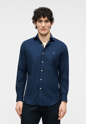 Jeune homme aux cheveux bouclés portant une chemise bleu marine à boutons avec un petit logo brodé sur la poitrine, sur un fond uni.