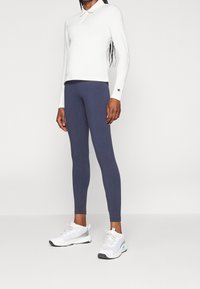 Vit långärmad polo med krage, kombinerad med marinblå leggings och vita sportskor med grå accenter och ljusblå detaljer.