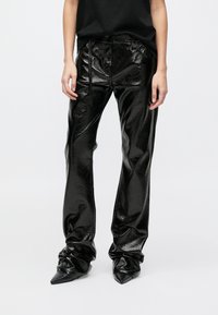 Μαύρο παντελόνι από δερμάτινο λάκα με γυαλιστερή υφή, χαλαρή εφαρμογή, μέση με κορδόνι και σούρες στα μανσέτα. Μαύρα μυτερά τακούνια.