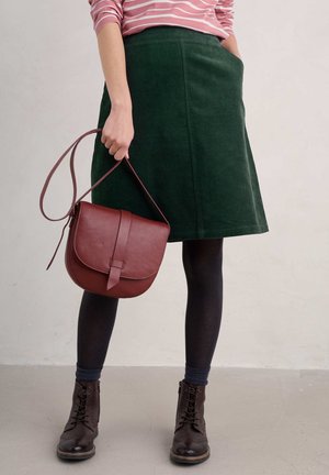 Femme portant une jupe vert foncé, des collants noirs, des bottes marron à lacets et un haut rose à rayures, tenant un sac à bandoulière en cuir marron.