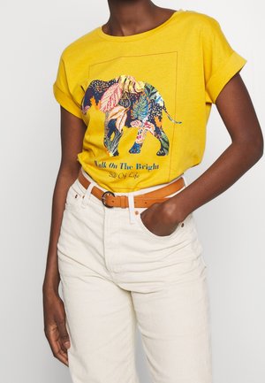 Personne portant un t-shirt jaune avec un éléphant à motifs multicolores et le texte "Walk On The Bright Side Of Life,", accompagné d'un pantalon blanc et d'une ceinture beige.
