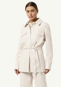 Jasno beżowa koszula-marynarka z paskiem, z dwiema przednimi kieszeniami, klasycznym kołnierzykiem oraz gładką, błyszczącą fakturą. Długie rękawy i dopasowany krój.