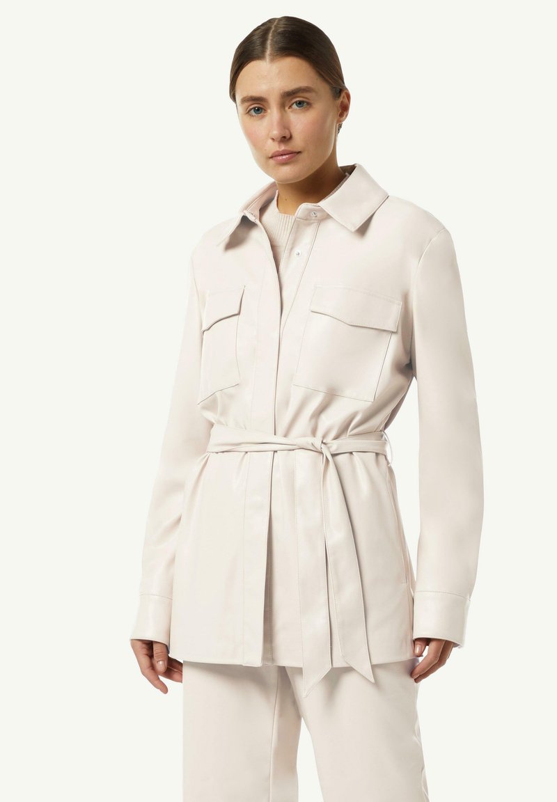 Jasno beżowa koszula-marynarka z paskiem, z dwiema przednimi kieszeniami, klasycznym kołnierzykiem oraz gładką, błyszczącą fakturą. Długie rękawy i dopasowany krój.