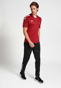 Rode sportieve shirt met korte mouwen en witte chevronaccenten op de schouders, gecombineerd met zwarte broek met witte strepen.