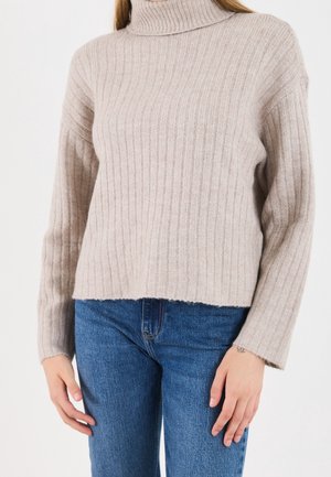 Personne portant un pull à col roulé côtelé beige et un jean bleu, debout devant un fond blanc uni.