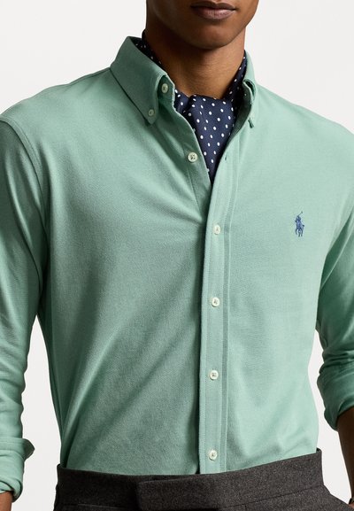 Camisa verde menta abotoada com um tecido texturizado, apresentando um pequeno logotipo azul e um colarinho abotoado. Embaixo, há um lenço azul marinho com bolinhas.
