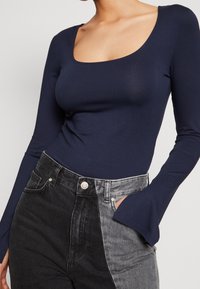 Marinblå långärmad bodysuit med rundad halsringning, parat med högmidjade svarta och grå färgblockerade jeans med en sidficka.