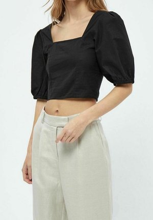 Femme portant un haut court noir à encolure carrée avec des manches bouffantes et un pantalon taille haute beige clair, debout devant un fond uni.