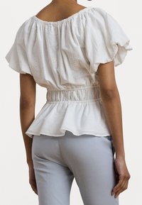 Vit bomullsblus med elastisk midja, puffärmar, broderie anglaise-detaljer och en peplumslut. Bärs med ljusblå byxor.