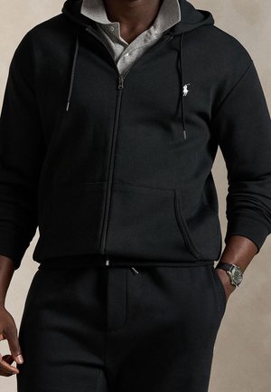 Muž v černé mikině na zip přes šedou košili s límečkem, v černých kalhotách a s černými hodinkami na zápěstí, s rukou v kapse na jednoduchém pozadí.