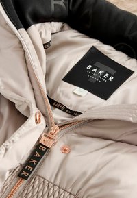 Chaqueta puffer de color beige claro con acabado satinado, forro interior negro, cremallera en tono dorado rosado y la marca 'BAKER' en la etiqueta y el tirador de la cremallera.
