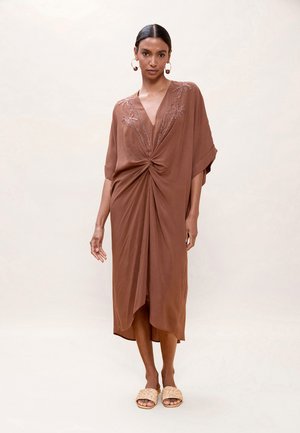 Mujer con un vestido midi marrón con escote en V y detalle torcido en el frente, bordado floral, combinado con sandalias beige de tela tejida y grandes pendientes de aro.