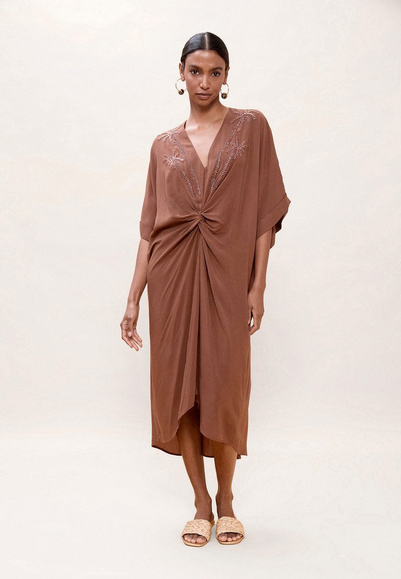 Femme portant une robe midi marron avec un col en V et un détail torsadé à l'avant, ornée de broderies florales, associée à des sandales beige tressées sans lacets et de grandes boucles d'oreilles.