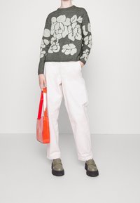 Personne portant un pull gris avec des motifs floraux blancs, un pantalon blanc à jambes larges, des mocassins verts massifs et tenant un sac à main orange vif.