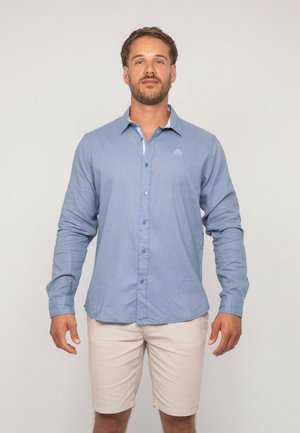 Homme debout, face à l'avant, portant une chemise à boutons bleu clair et un short beige, sur un fond uni.
