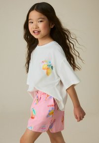 Jong meisje met lang donker haar, een wit t-shirt met citroenen en roze korte broek met kleurrijke zomerprints, loopt zijwaarts.