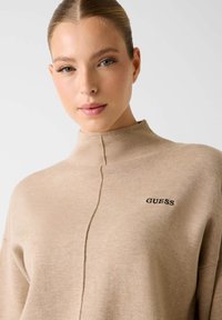Beige coltrui gemaakt van zacht gebreide stof, met een naaddetail aan de voorkant en het "GUESS" logo in zwarte borduring op de borst.