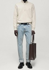 Persona vistiendo un suéter crema con media cremallera, jeans azul claro con puños, cinturón negro, zapatos de cuero negros y sosteniendo un bolso tote de cuero marrón oscuro.