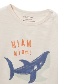 T-shirt en coton de couleur crème avec un graphisme de requin bleu et le texte orange "MIAM miam!" sur le devant. Col rond et tissu doux.