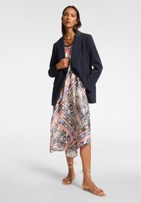 Marineblazer über einem gemusterten Kleid in Rosa, Blau und Creme. Das Kleid hat ein fließendes Design und einen asymmetrischen Saum. Goldene Sandalen mit Knöchelbindungen.