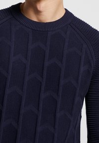 Marinblå stickad tröja med en texturerad, geometrisk mönster på framsidan, ribbad krage och ärmsömmar, med en figursydd design.