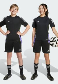 Due bambini in maglie e pantaloni sportivi neri Adidas con accenti laterali verdi e viola. Indossano calze da calcio nere e scarpini, uno tiene un pallone da calcio.