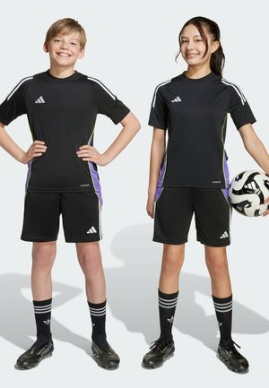 Due bambini in maglie e pantaloni sportivi neri Adidas con accenti laterali verdi e viola. Indossano calze da calcio nere e scarpini, uno tiene un pallone da calcio.
