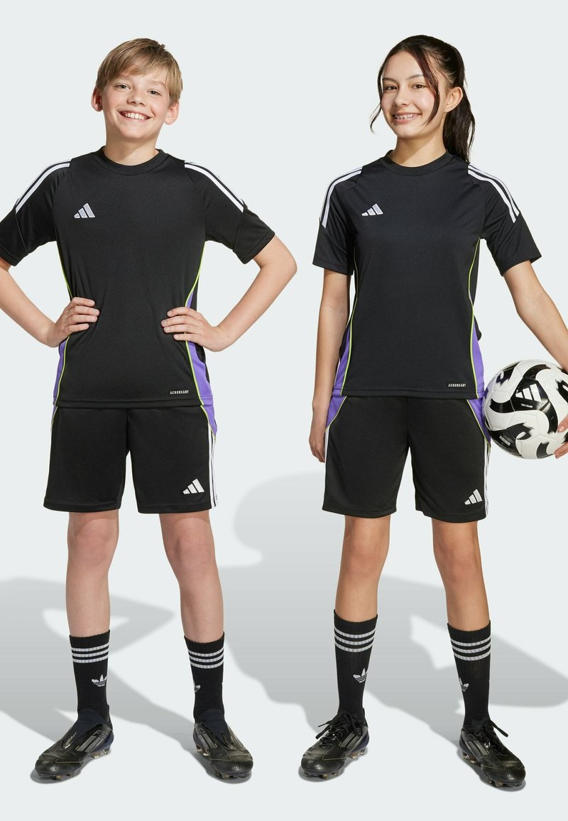 Two children in black Adidas athletic jerseys and shorts with green and purple side accents. They wear black soccer socks and cleats, one holding a soccer ball.