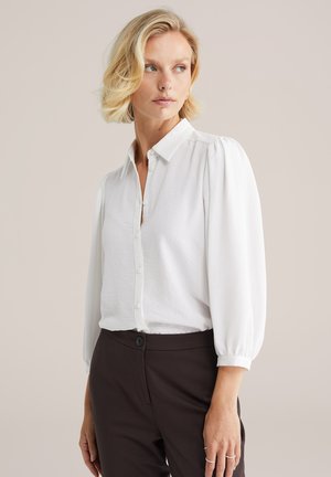 White blouse with a buttoned placket, collared neckline, and puff sleeves. Textured fabric and a relaxed fit. Paired with dark trousers.