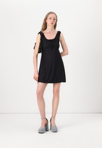 Vestido preto sem mangas com ombros franzidos, cintura ajustada e tecido texturizado. Combinado com sapatos de salto cinza com tiras para os tornozelos.