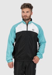 Windbreaker jacket in zwart, wit en turquoise, met een ritssluiting aan de voorkant, elastische manchetten en een logo op de linkerkant van de borst. Lichtgewicht materiaal.