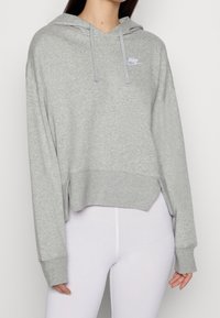 Grå kortärmad hoodie i mjukt tyg med en frontficka, vida ärmar och en vit Nike-logotyp på vänster bröst.