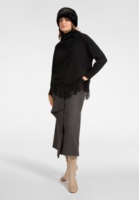 Pull en tricot noir avec détail à franges en bas, associé à une jupe portefeuille grise et des bottes beiges jusqu'aux genoux. Modèle portant un chapeau en fourrure noir.