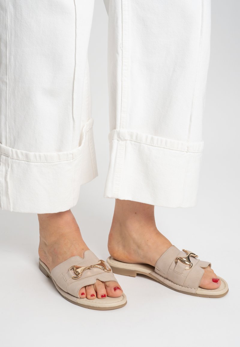 Béžové pantofle s texturovaným svrchním dílem, zlatým řetízkovým detailem a polstrovanou podrážkou, spojené s bílými kalhotami širokých nohavic.