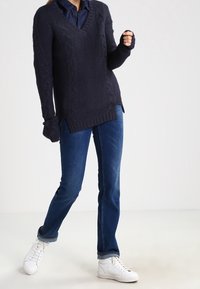 Marinblå kabelstickad tröja med V-ringning och långa ärmar, kombinerad med mörkblå jeans och vita sneakers, med en avslappnad passform.