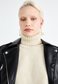 Cream ribbed knit turtleneck under a black leather jacket with silver hardware. Large hoop earrings and smooth skin texture visible.