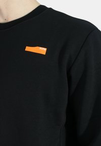 Felpa nera con scollatura a girocollo presenta un piccolo logo rettangolare arancione sul lato sinistro. Il tessuto appare liscio e strutturato.