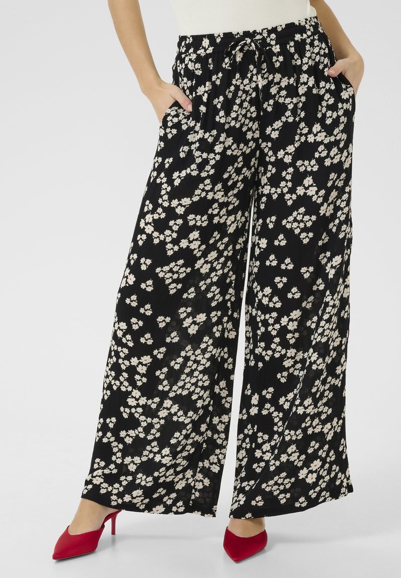 Femme portant un pantalon noir à jambes larges avec un imprimé floral blanc et des chaussures rouges à talons hauts, mains dans les poches, debout devant un fond uni.