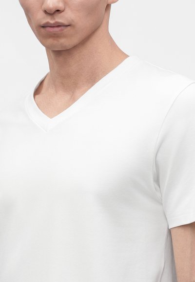 Gros plan d'un homme portant un t-shirt blanc uni à col en V et manches courtes sur un fond gris clair.