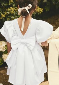 Robe blanche avec des manches bouffantes, décolleté en V profond et grand nœud à la taille. Tissu lisse avec une fermeture à bouton, placée devant une verdure.
