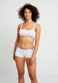 Νέα γυναίκα που στέκεται, φορώντας λευκή τοπ με διπλές τιράντες και λευκό εσώρουχο boyshort, μπροστά από ένα απλό ανοιχτό γκρι φόντο.