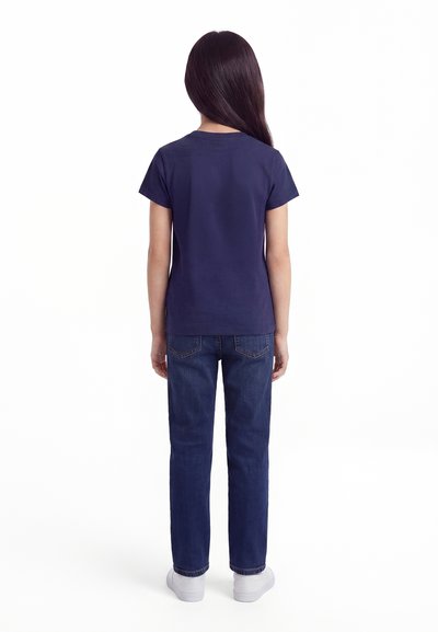 Copil cu păr lung și întunecat, stând cu fața spre spate, purtând un tricou bleumarin cu mânecă scurtă, blugi albaștri și adidași albi pe un fundal alb.