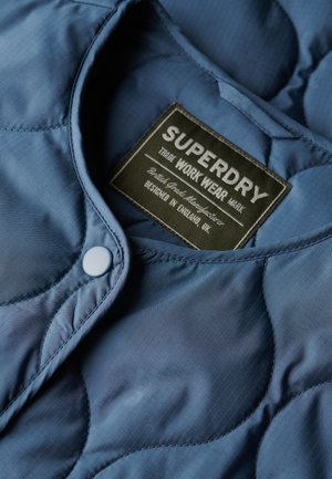 Gros plan d'une veste de travail matelassée bleue avec un bouton-pression et une étiquette de la marque Superdry à l'intérieur du col.