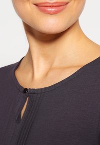 Ciemnoszara bluzka z okrągłym dekoltem, plisowanymi detalami oraz zapięciem na guzik przy dekolcie. Materiał wydaje się miękki i gładki.