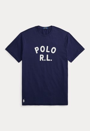Muž stojaci s rukami v kapsách, oblečený v tmavomodré tričko "POLO R.L.", biele nohavice, hnedý opasok, náramkové hodinky a tmavomodré tenisky.