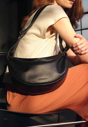 Borsa a spalla in pelle nera con una superficie liscia e un design curvo. Presenta una lunga tracolla e un branding sobrio. Indossata con una maglietta crema e pantaloni arancioni.
