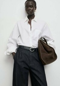 Camisa blanca de botón con mangas enrolladas, pantalones oscuros de cintura alta, cinturón negro con hebilla circular y bolso de hombro marrón.