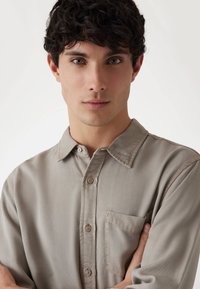 Camisa de botones color beige claro con cuello, un bolsillo en el pecho y tela suave y texturizada. Los botones son de un tono ligeramente más oscuro.
