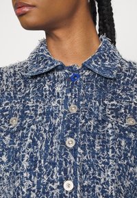 Zbliżenie osoby noszącej niebiesko-biały wełniany żakiet o teksturze, z srebrnymi guzikami i kołnierzem, widoczna szyja i dolna część twarzy.